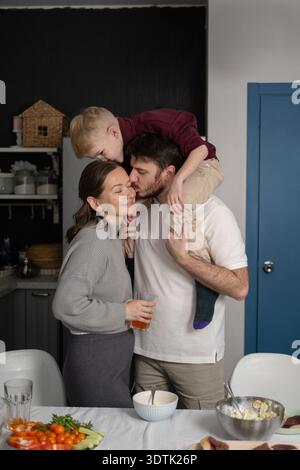 Una famiglia amorevole condivide un momento tenero in cucina, mentre il padre bacia la madre tenendo il bambino sulle spalle, circondato da fresco Foto Stock