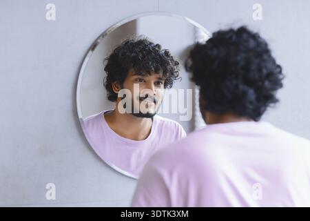 Young Man guarda nello specchio e sorride durante la routine mattutina nella Bright Room Foto Stock