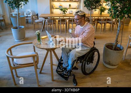 Allegro positivo disabile uomo in sedia a rotelle che lavora con un computer portatile e beve caffè in un accogliente caffè. Foto Stock