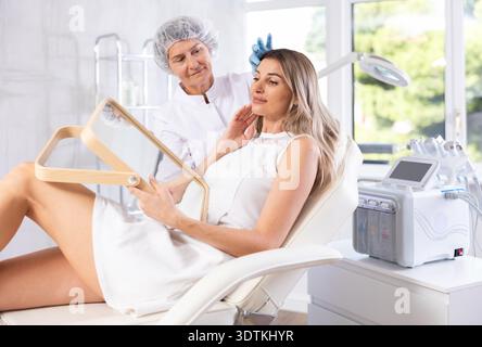 Giovane donna bionda soddisfatta che guarda nello specchio dopo un intervento di chirurgia plastica. Consultazione con chirurgo professionista Foto Stock
