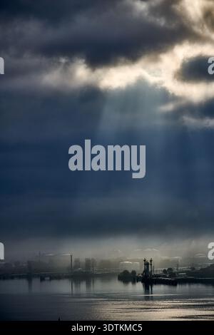 Una fotografia suggestiva e suggestiva che cattura pozzi di luce solare che penetrano attraverso nuvole scure e pesanti, illuminando un paesaggio industriale lontano Foto Stock