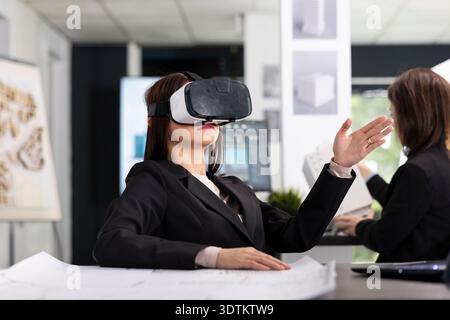 Un'architetto professionista si siede in ufficio indossando un visore VR ed esaminando le planimetrie per il progetto di un edificio. Donna che esamina i layout 3D e il flusso di lavoro di pianificazione, utilizzando strumenti digitali e immersivi. Foto Stock