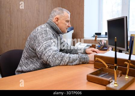 L'anziano uomo caucasico con i capelli grigi si siede a una scrivania, concentrato e triste, al lavoro su un computer. Foto Stock
