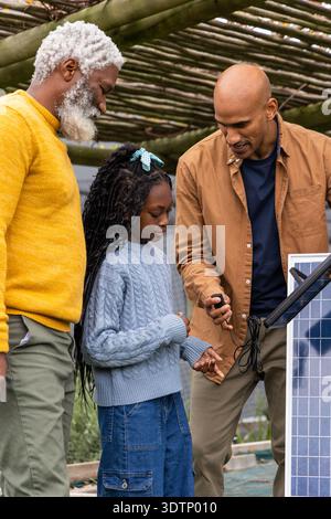 Famiglia eterogenea uomo anziano e bambino ragazza che guarda l'uomo che prova i cavi del pannello solare sotto il pergolato di legno Foto Stock