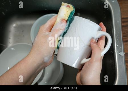 Primo piano delle mani che puliscono una tazza di ceramica bianca con una spugna saponata in un lavandino in acciaio inox. La schiuma copre la superficie durante la rimozione dei residui. Foto Stock