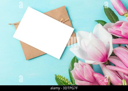Fiori di magnolia rosa e foglie verdi con biglietto d'auguri bianco e busta su sfondo turchese. Mockup floreale con spazio di copia. Matrimonio Foto Stock
