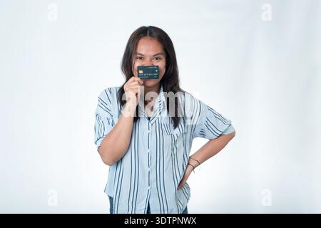 Donna indonesiana con una camicia a righe blu che tiene una carta di credito nera di alta qualità davanti al viso, coprendo la bocca. Occhi sorridenti e posa sicura Foto Stock
