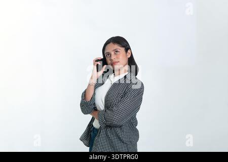 Ritratto di una donna indonesiana con una camicia a quadri che tiene un telefono all'orecchio. Sta guardando verso l'alto con un'espressione premurosa. Isolato su bianco. Foto Stock