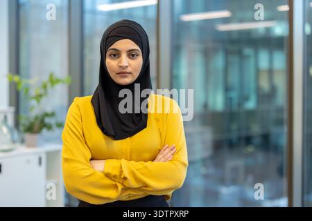 Donna d'affari musulmana che indossa un hijab e una camicia gialla, in piedi con fiducia con le braccia piegate in un ufficio moderno, rappresentando professionalità, empowerment e successo nella sua carriera Foto Stock
