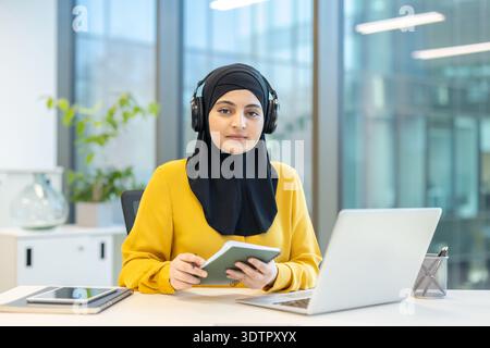 Donna musulmana che indossa un hijab nero e una camicia gialla, seduta a una scrivania con le cuffie, guarda la fotocamera mentre tiene in mano un notebook e lavora con un computer portatile Foto Stock