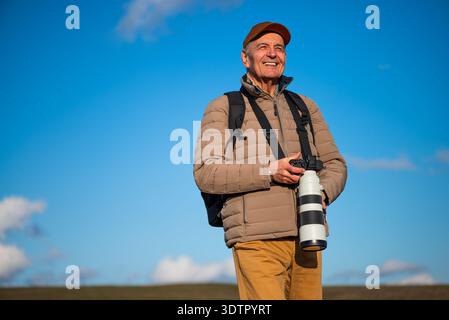 Un uomo anziano con fotocamera digitale e teleobiettivo esplora la natura, concentrandosi sullo scatto perfetto. Foto Stock