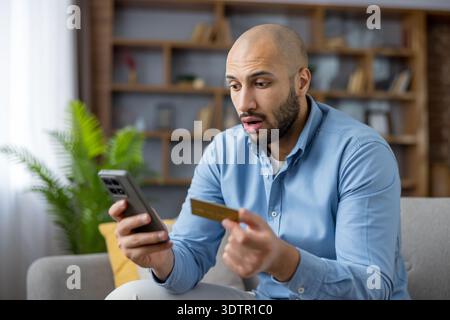 Giovane che sta vivendo uno shock finanziario e si preoccupa mentre controlla un pagamento online o un'applicazione bancaria sul suo smartphone, tenendo in mano una carta di credito oro Foto Stock