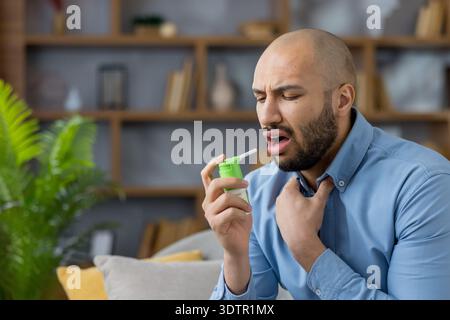 Giovane uomo nero al chiuso che applica spray alla gola per aprire la bocca mentre tocca il collo, mostrando disagio per mal di gola o influenza mentre cerca sollievo e cura di sé a casa Foto Stock