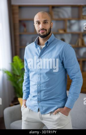 Uomo calvo con barba che indossa una camicia azzurra e pantaloni beige, in piedi con le mani in tasca, guardando direttamente la fotocamera in un ambiente moderno di casa o ufficio Foto Stock
