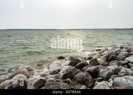 La fredda luce invernale si riflette sul mare mentre le rocce ghiacciate costeggiano il litorale, creando una tranquilla ma aspra scena costiera. Foto Stock