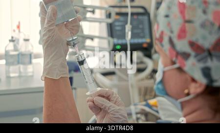 Infermiere che aspira il farmaco nella siringa in terapia intensiva. L'operatore sanitario con guanti riempie una siringa da un flaconcino accanto ai monitor per terapia intensiva, preparando la sterilizzazione Foto Stock