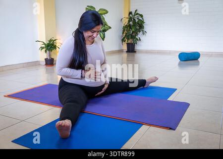 Donna incinta che esercita yoga, tiene la pancia e allunga le gambe in uno studio Foto Stock