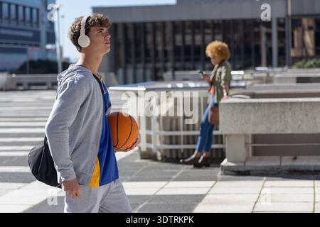 Adolescente in maglia che cammina in plaza tenendo il basket mentre una donna afroamericana usa il telefono Foto Stock