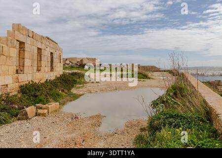 La vista dei muri in pietra invecchiata incontra l'orizzonte sotto un vasto cielo, riflesso in una pozzanghera ancora intatta, con texture contrastanti e sfumature naturali, il-Mellieħa, Malta. Foto Stock