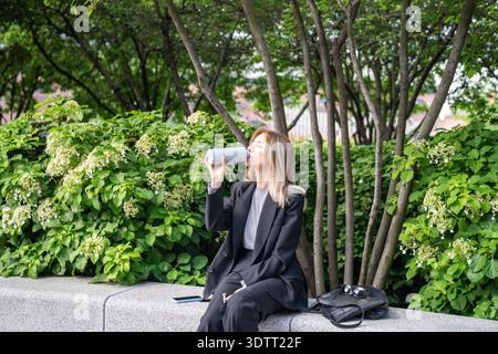 Una donna d'affari rilassata riposa dopo una giornata in ufficio nel parco, gode del tempo e beve acqua dalla bottiglia. Foto Stock