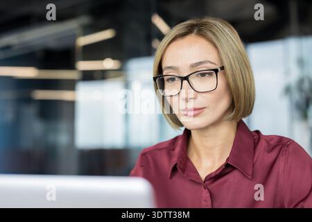 Manager professionista donna che indossa occhiali da vista e una camicia marrone, concentrandosi con attenzione sullo schermo del suo notebook in un ambiente d'ufficio moderno, che rappresenta Foto Stock
