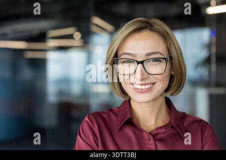 Donna d'affari sicura di sé con un'espressione positiva, indossa occhiali da vista e una camicia marrone, che rappresenta professionalità e leadership in un look moderno Foto Stock