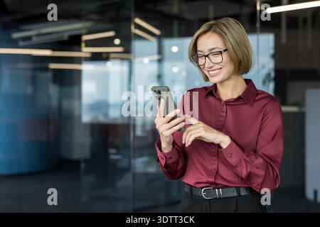 Donna d'affari professionista sorridente mentre utilizza attivamente uno smartphone, si occupa di comunicazione digitale, lavoro, networking o gestione del suo frenetico sch Foto Stock