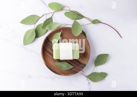 Sbucciare sapone verde alle erbe con copertura di carta vuota e rametti di eucalipto aromatizzati freschi su un piatto di legno su fondo di marmo bianco. Bagno per la cura del corpo o. Foto Stock