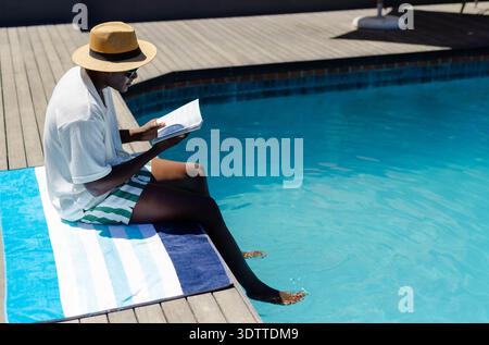 Libro cartaceo aperto che poggia su un asciugamano blu e bianco a bordo piscina con cappello di paglia e occhiali da sole Foto Stock