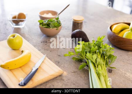 Mazzo di sedano appoggiato su un piano di lavoro in pietra, con tagliere che contiene banana, mela verde, coltello Foto Stock