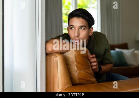 Uomo afroamericano appoggiato sul retro del divano in pelle caramellata che guarda alla finestra indossando un orologio da polso scuro Foto Stock