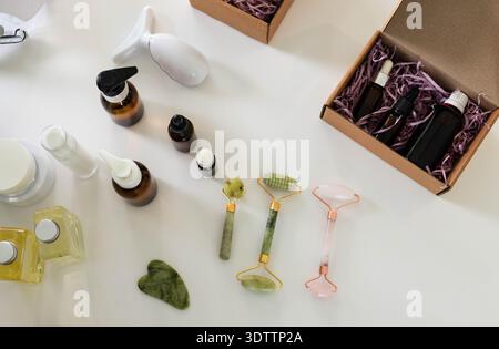 Tre rulli facciali e pietra di gua Sha poggiati su un piano bianco con bottiglie per la cura della pelle nelle vicinanze Foto Stock