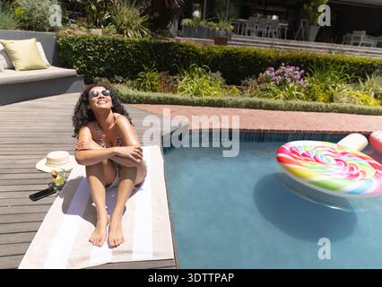 Donna afroamericana in costume da bagno a motivi geometrici seduta a bordo piscina su asciugamano con cappello di paglia e telefono Foto Stock