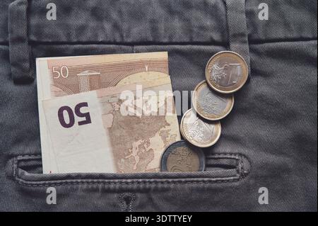 banconota in euro parzialmente visibile nella tasca posteriore di un paio di pantaloni, con monete in euro disposte in una riga ordinata accanto. Concetto di denaro, denaro, risparmio Foto Stock