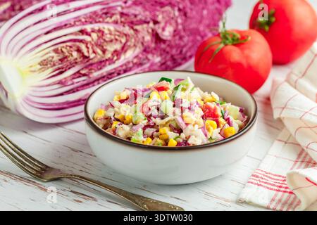 Deliziosa insalata di cavolo rosso alla peeling con pomodori, cetrioli, mais, cipolle e bastoncini di granchio in un piatto bianco su un tavolo di legno. Cena gourmet leggera. Se Foto Stock