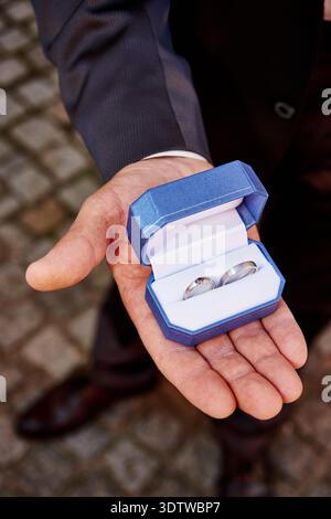 Uno scatto ravvicinato ad angolo alto cattura la mano di un uomo, vestito con una giacca scura e una camicia bianca, che regge un anello aperto in velluto blu reale Foto Stock