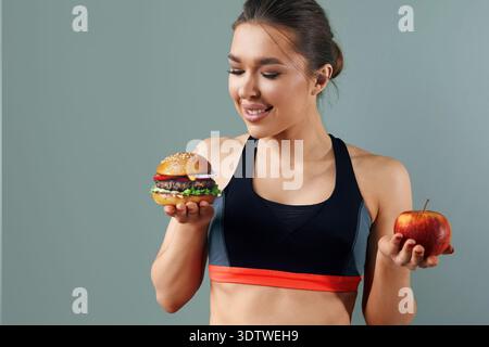Una giovane donna in abbigliamento sportivo si erge su uno sfondo grigio, tenendo una mela in una mano e un hamburger nell'altra, contemplando la sua scelta di cibo Foto Stock