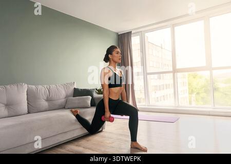 Una donna in forma si affonda con un manubrio in mano in un elegante salotto. Il sole splende attraverso grandi finestre, creando un luminoso e motivato Foto Stock