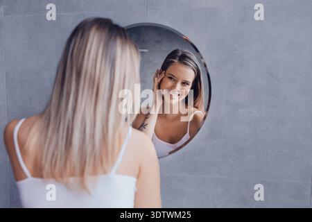 La donna ammira il suo riflesso mentre sorride in uno specchio rotondo in un bagno moderno Foto Stock