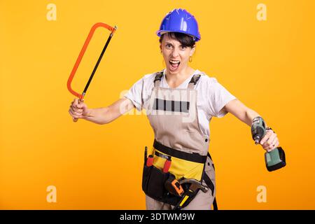 Repairwoman grida con entusiasmo, tenendo in mano sega a mano e trapano elettrico. Una donna che rinnova la sua attività non vede l'ora di fornire servizi di tuttofare e di migliorare la casa con gli attrezzi da costruzione a portata di mano. Foto Stock