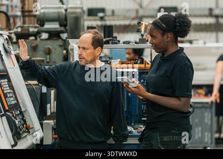 Lavoratrice che prende appunti mentre la collega spiega le parti della macchina in fabbrica Foto Stock