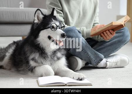 Giovane seduto sul pavimento e leggendo un libro con un simpatico cane husky in occhiali da vista a casa Foto Stock