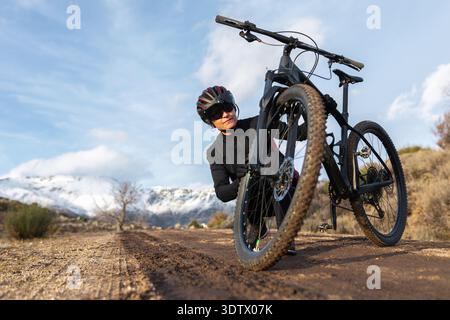 Una donna che indossa un casco ispeziona lo pneumatico della sua mountain bike su un aspro sentiero sterrato. Montagne innevate si stagliano maestosamente sullo sfondo e Foto Stock