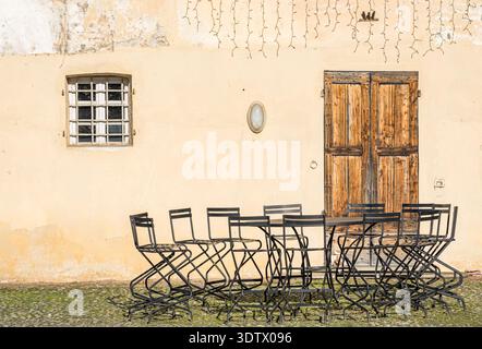 Un caffe' esterno vuoto con sedie di metallo e un tavolo di legno adagiato su di una vecchia parete di stucco, trasuda fascino rustico e invitante tranquillita' su di un soleggiato giardino Foto Stock