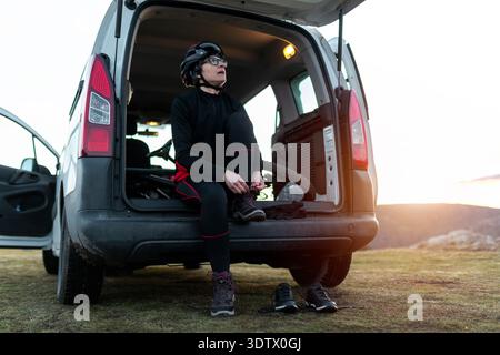 Una donna siede sul retro di un furgone parcheggiato all'alba, legando le sue scarpe. Pronta per un'avventura in mountain bike, indossa un casco e guarda verso l'alto Foto Stock