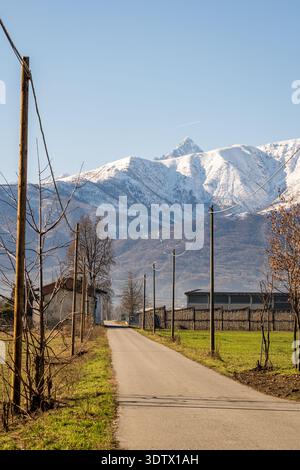 Una tranquilla strada rurale in Piemonte conduce verso il Monviso innevato sotto un cielo azzurro, fiancheggiato da campi e alberi, creando un'immagine Foto Stock