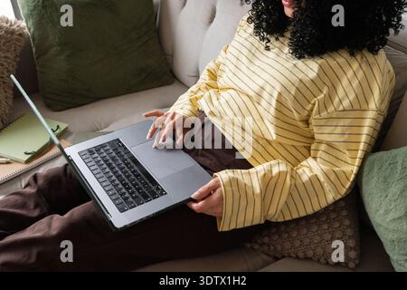 Una donna con un maglione giallo a righe lavora sul suo laptop mentre è seduta comodamente su un divano. Si occupa di lavoro a distanza dall'ambiente accogliente Foto Stock