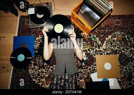 Giovane donna che copre il viso con un disco in vinile mentre si sdraia su un tappeto Foto Stock
