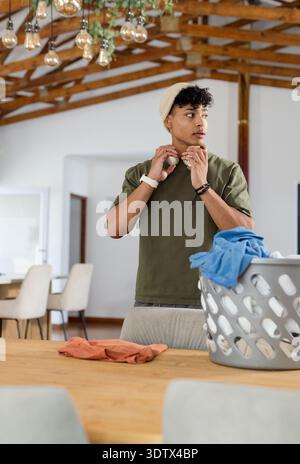 Uomo che regola le cuffie wireless dietro il tavolo da pranzo a casa, indossa un berretto vicino al cestino della biancheria Foto Stock