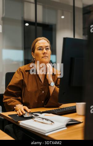 Donna d'affari matura e premurosa che lavora al computer mentre siede alla scrivania in un ufficio moderno Foto Stock